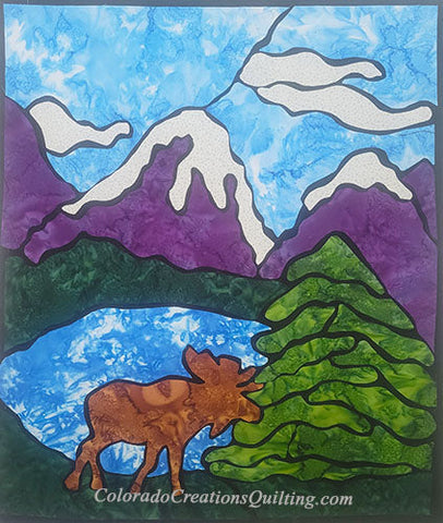 The quilt features a moose at a mountain lake surrounded by evergreen trees and puple mountains under blue sky with the look and feel of stained-glass.