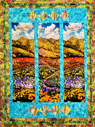 This quilt features a  meadow full of colorful flowers below  rolling hills and a bright blue cloudy sky.